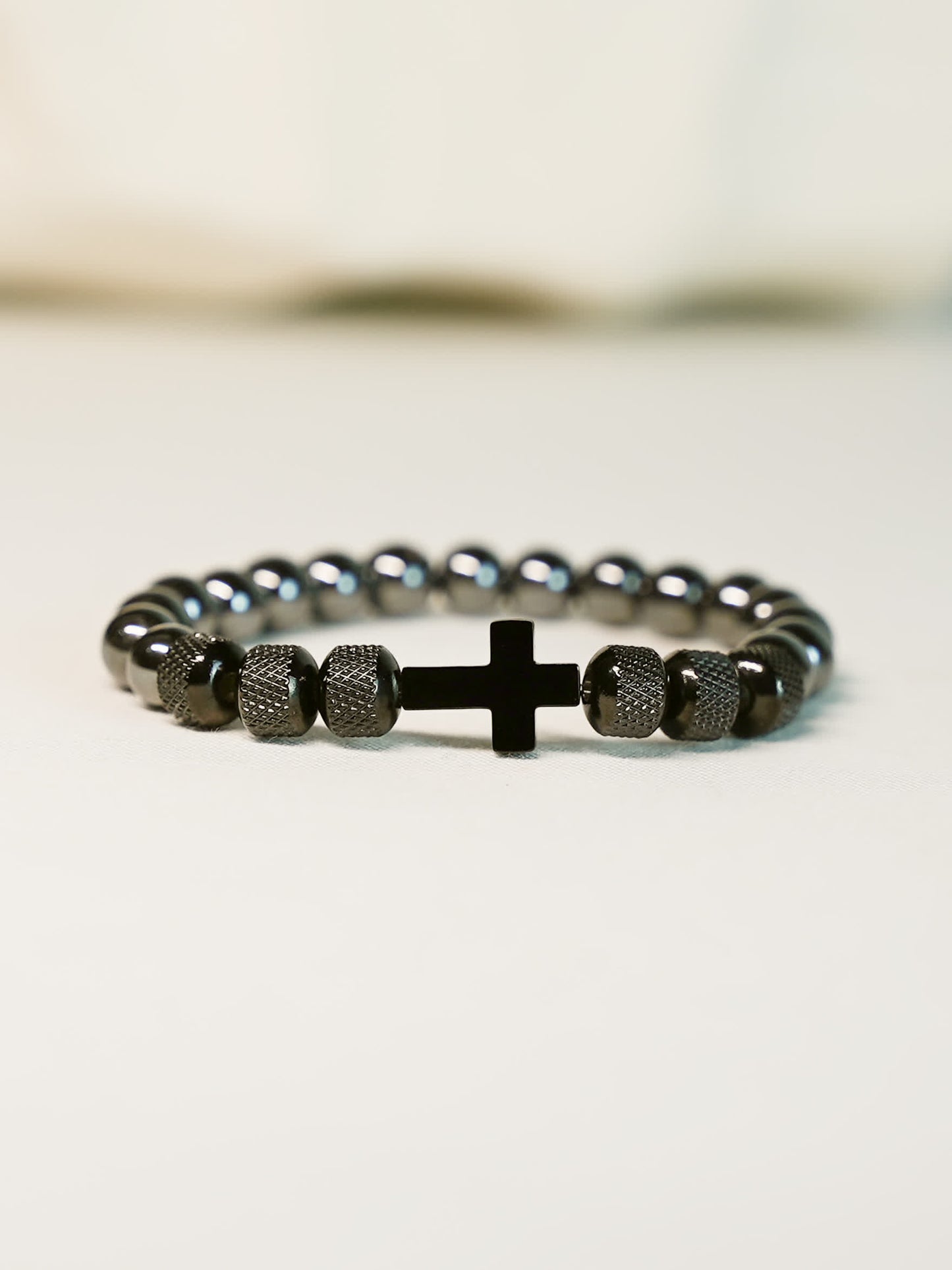 Side angle view of the Iron Grace Beaded Cross Bracelet resting on a cream-toned surface, emphasizing the modern design and stretch-fit structure.

