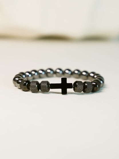 Side angle view of the Iron Grace Beaded Cross Bracelet resting on a cream-toned surface, emphasizing the modern design and stretch-fit structure.

