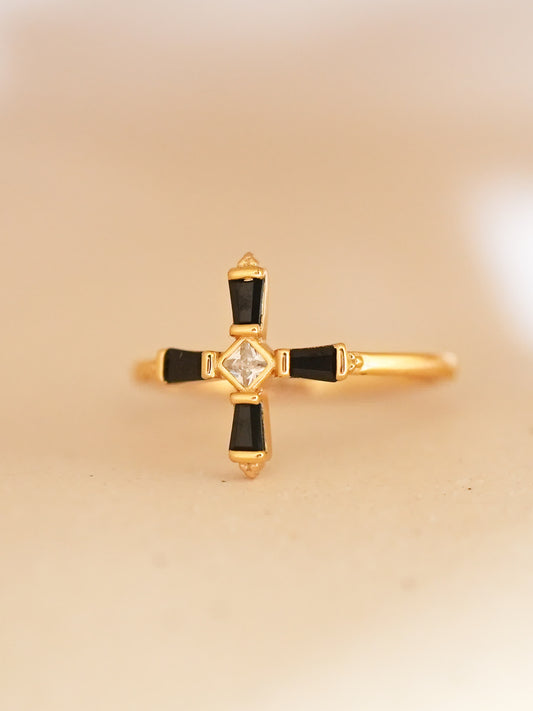 An angled photograph of the black and gold cross ring resting upright, showing its sleek band and symmetrical gemstone design in soft lighting.