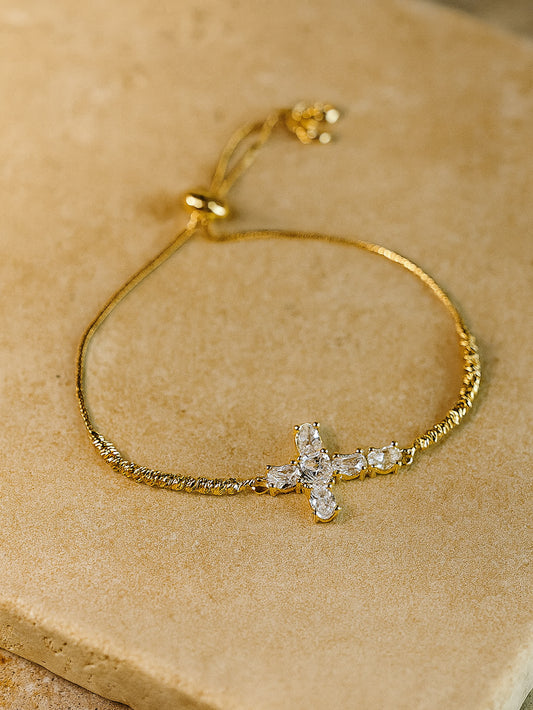 Elegant flat lay image of the Radiant Mercy Gold Cross Bracelet on a textured beige stone surface, featuring a sparkling zircon cross on a fine gold-toned band.