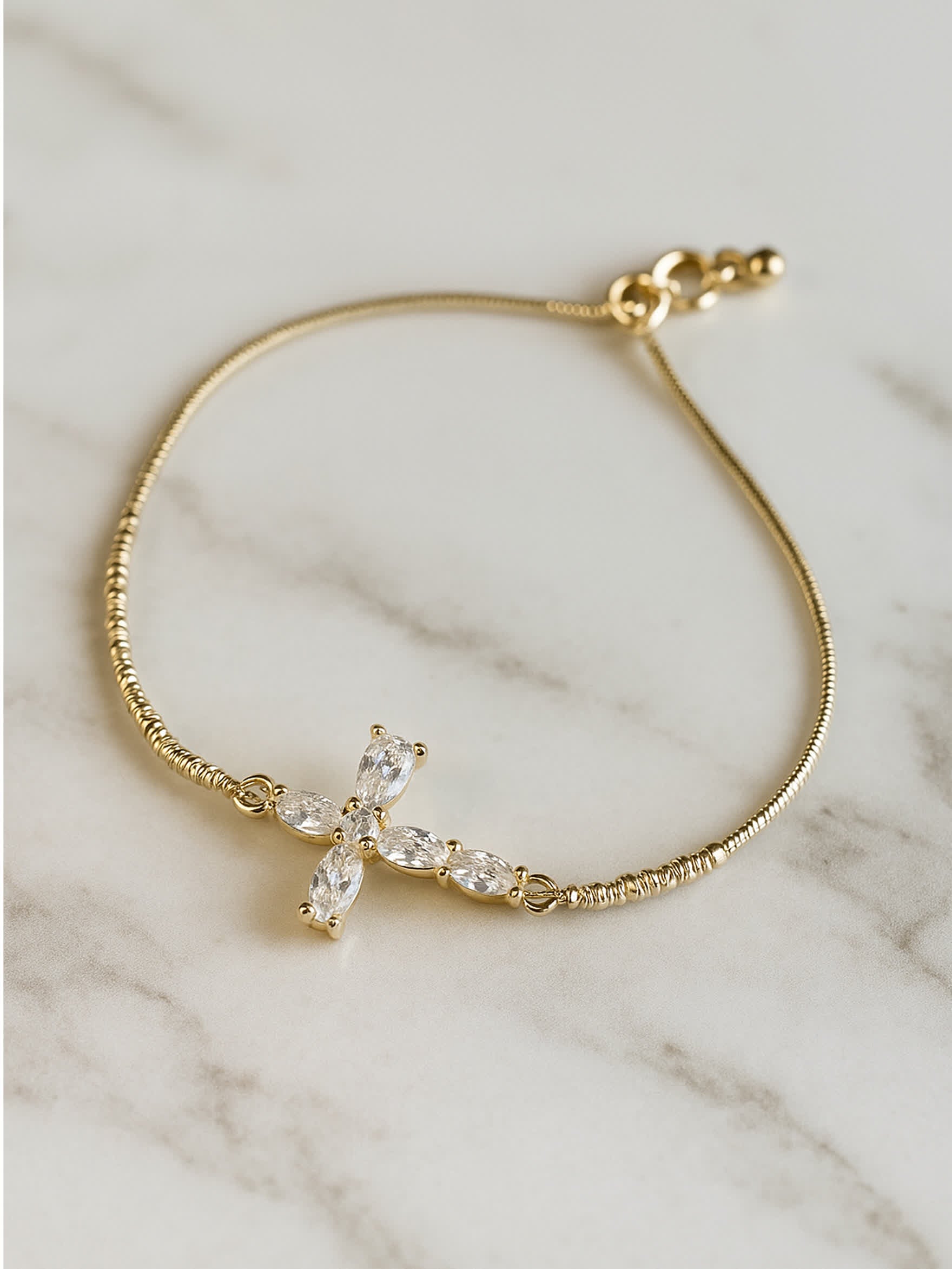 Top-down image of the Radiant Mercy Gold Cross Bracelet resting on a white marble surface, emphasizing the golden chain and glimmering cross centerpiece.