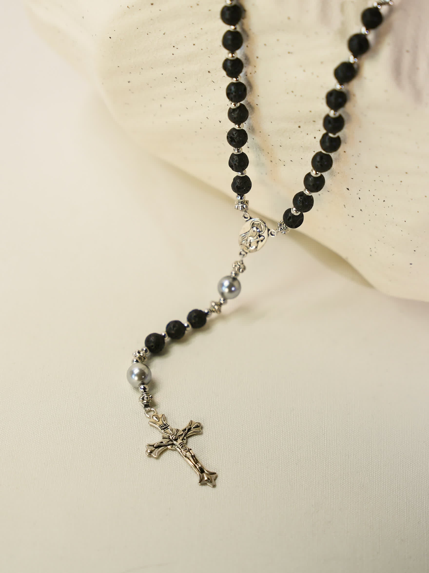 Volcanic rock rosary necklace with silver crucifix displayed on soft cream fabric