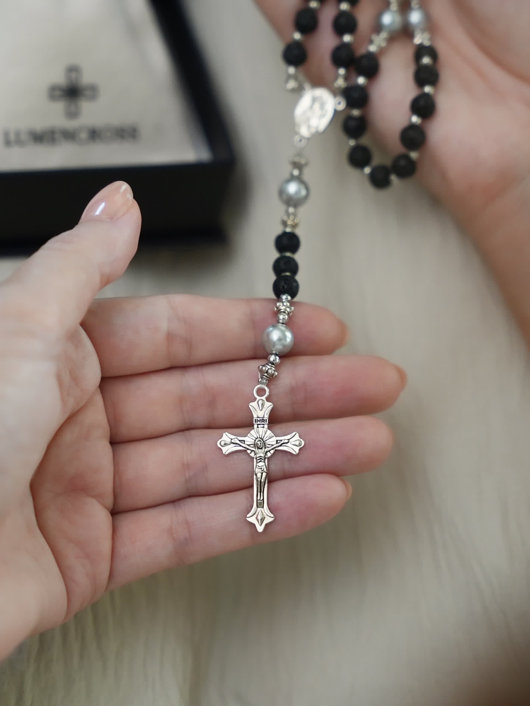 Volcanic lava rosary necklace with cross shown in hand next to Lumencross gift box