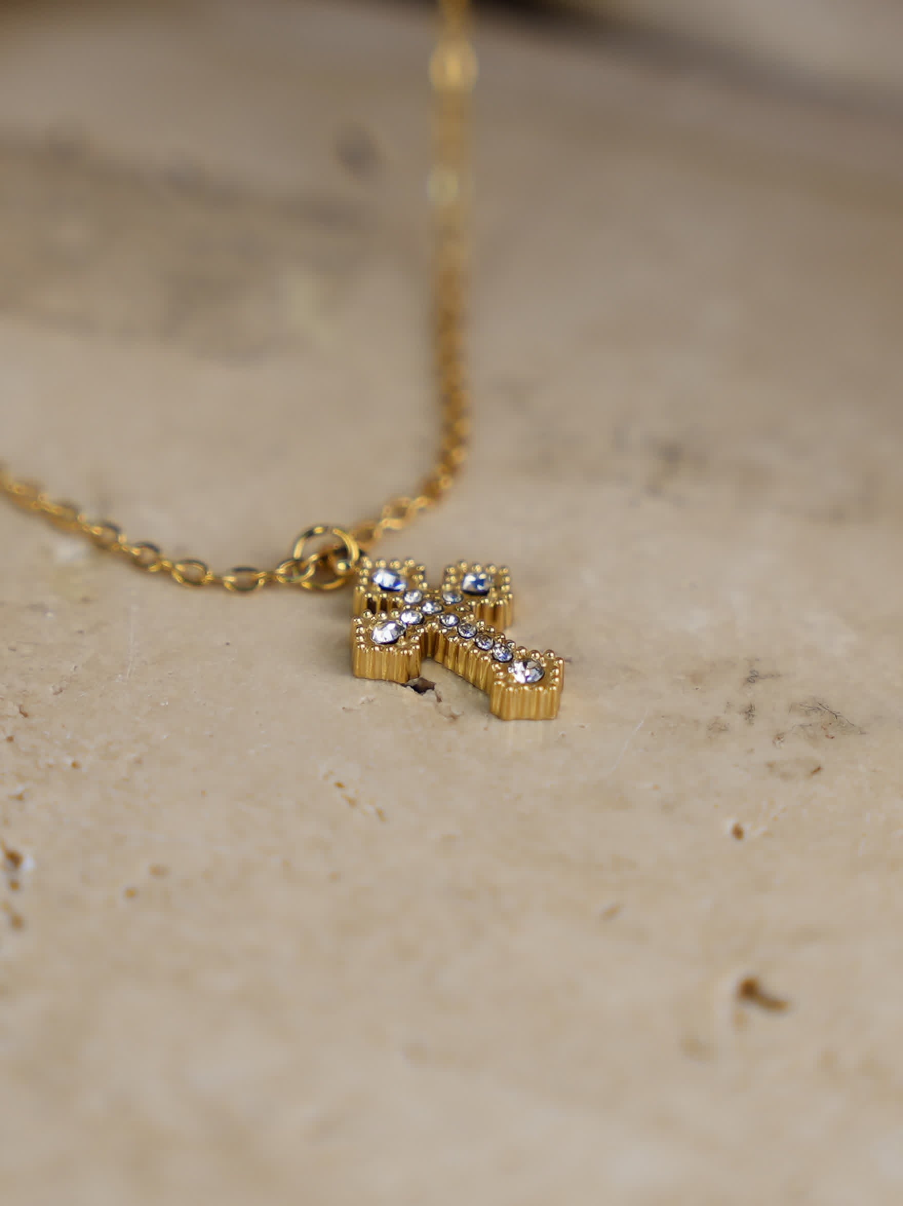 Small gold cross necklace with zircons laid flat on neutral stone surface