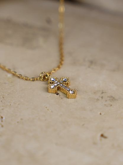 Small gold cross necklace with zircons laid flat on neutral stone surface