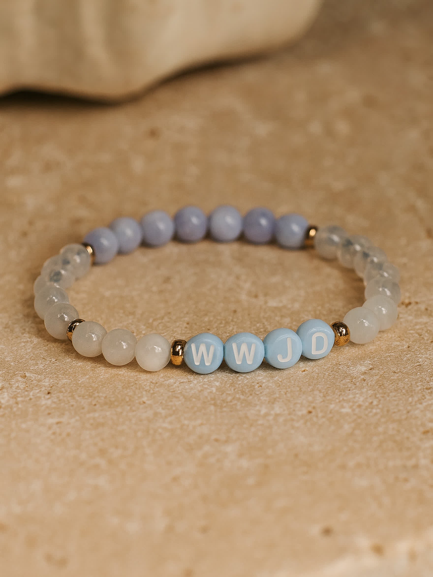 Flat lay product image of the Quiet Steps WWJD Bracelet on a stone surface, featuring natural gemstone beads and blue-and-white letter beads spelling WWJD with gold accents.