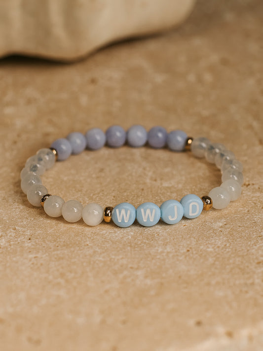 Flat lay product image of the Quiet Steps WWJD Bracelet on a stone surface, featuring natural gemstone beads and blue-and-white letter beads spelling WWJD with gold accents.