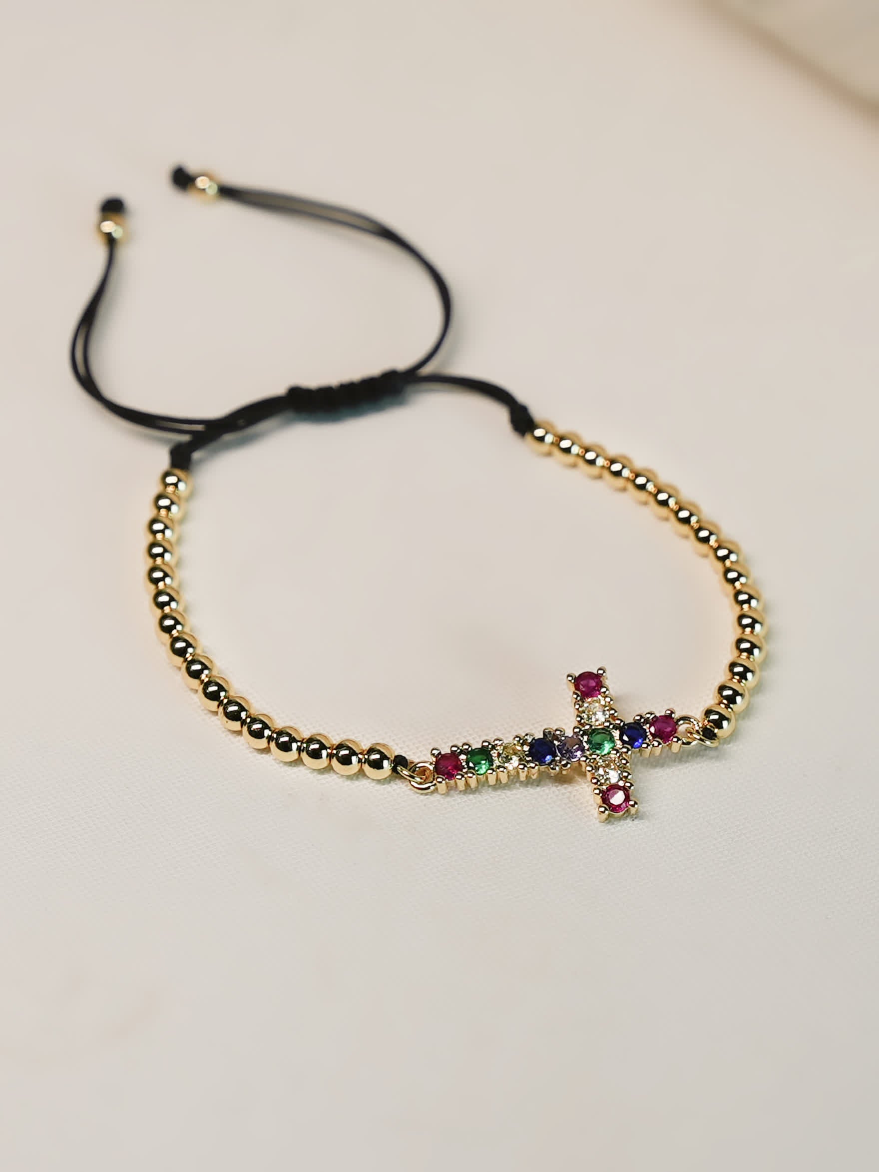 Top-down product photo of the Joy in Light Cross Bracelet on a soft neutral surface, showing the full bracelet shape, black pull-tie cord, and vibrant gemstone cross.