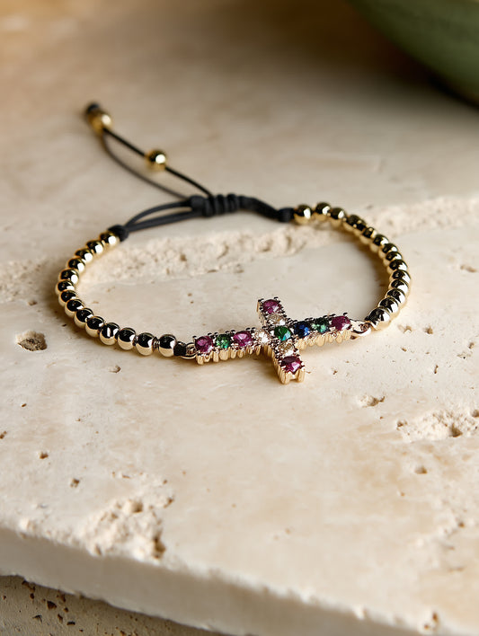 Front-facing image of the Joy in Light Cross Bracelet on a travertine surface, featuring gold-toned beads, a multicolor zircon cross, and an adjustable black cord.