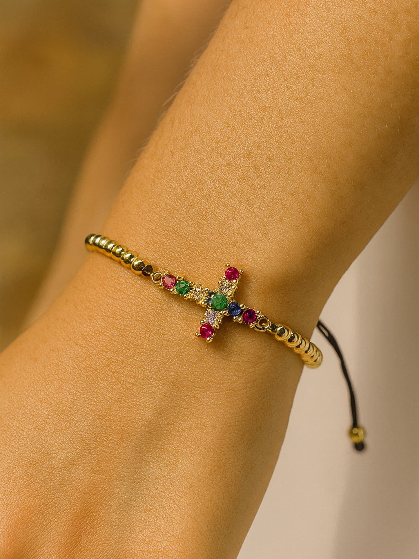 Lifestyle photo of the Joy in Light Cross Bracelet worn on a woman’s wrist, displaying the gold beaded band and rainbow-colored cross against warm natural lighting.