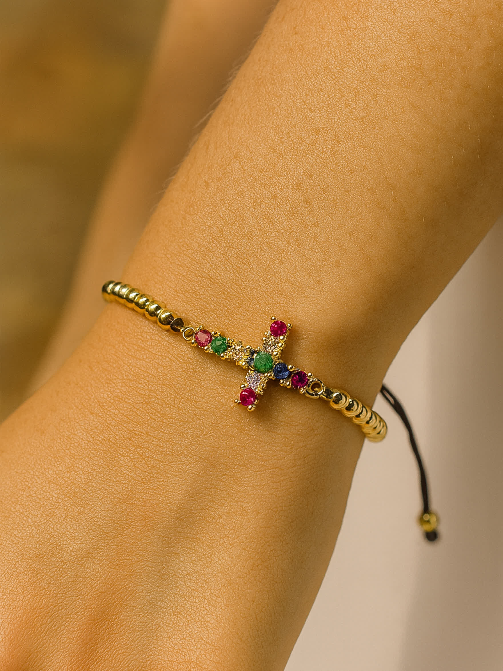 Lifestyle photo of the Joy in Light Cross Bracelet worn on a woman’s wrist, displaying the gold beaded band and rainbow-colored cross against warm natural lighting.
