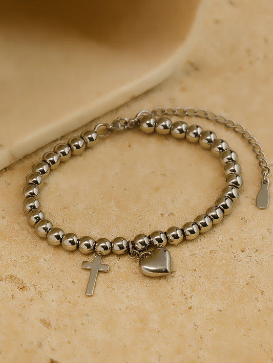 Flat lay photo of the Pray Through It Cross Charm Bracelet on a beige stone surface, featuring polished stainless steel beads with dangling cross and heart charms.