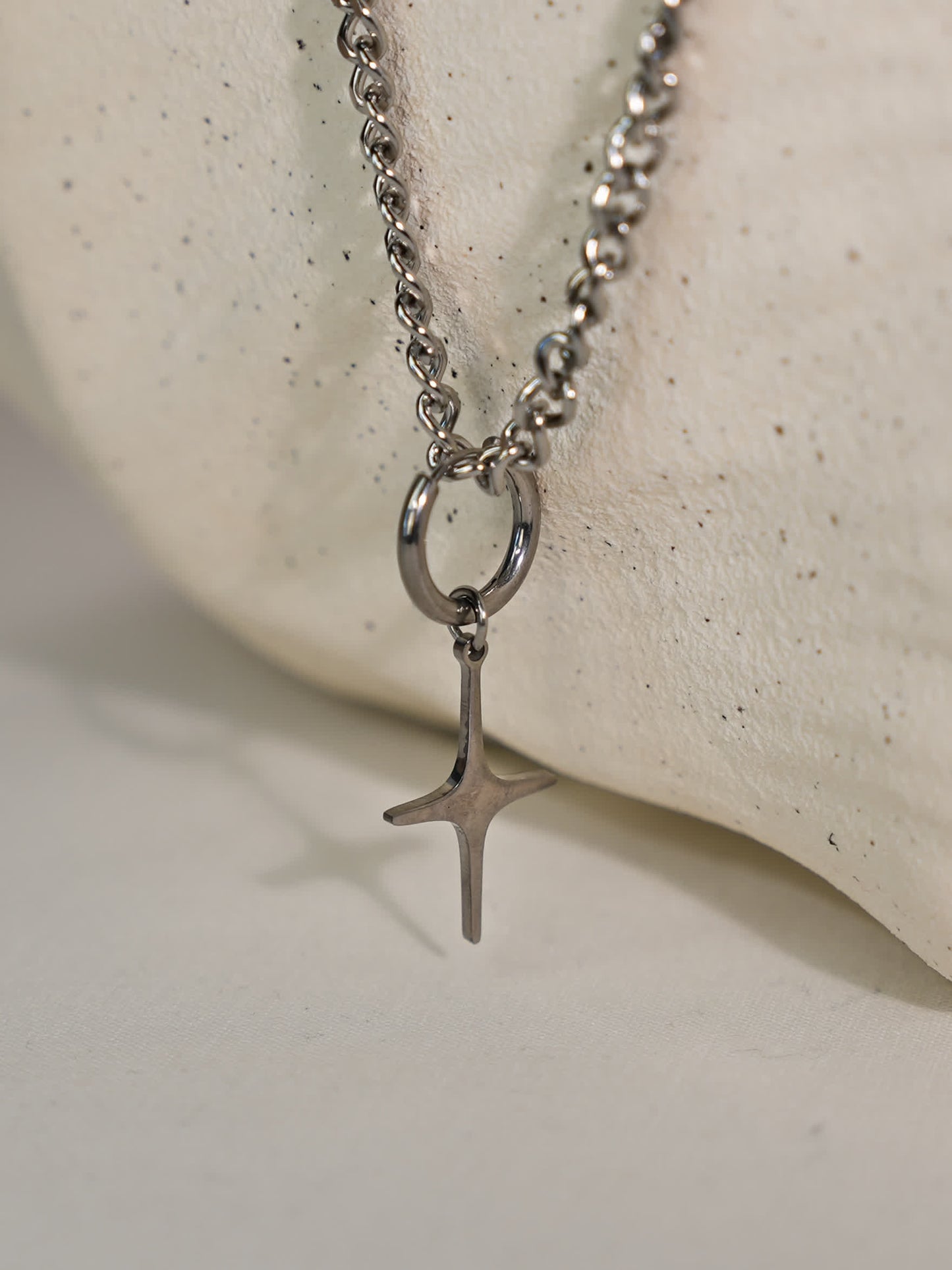 Brushed steel sculptural cross pendant hanging from bold chain, displayed on a curved white stone stand with soft lighting.