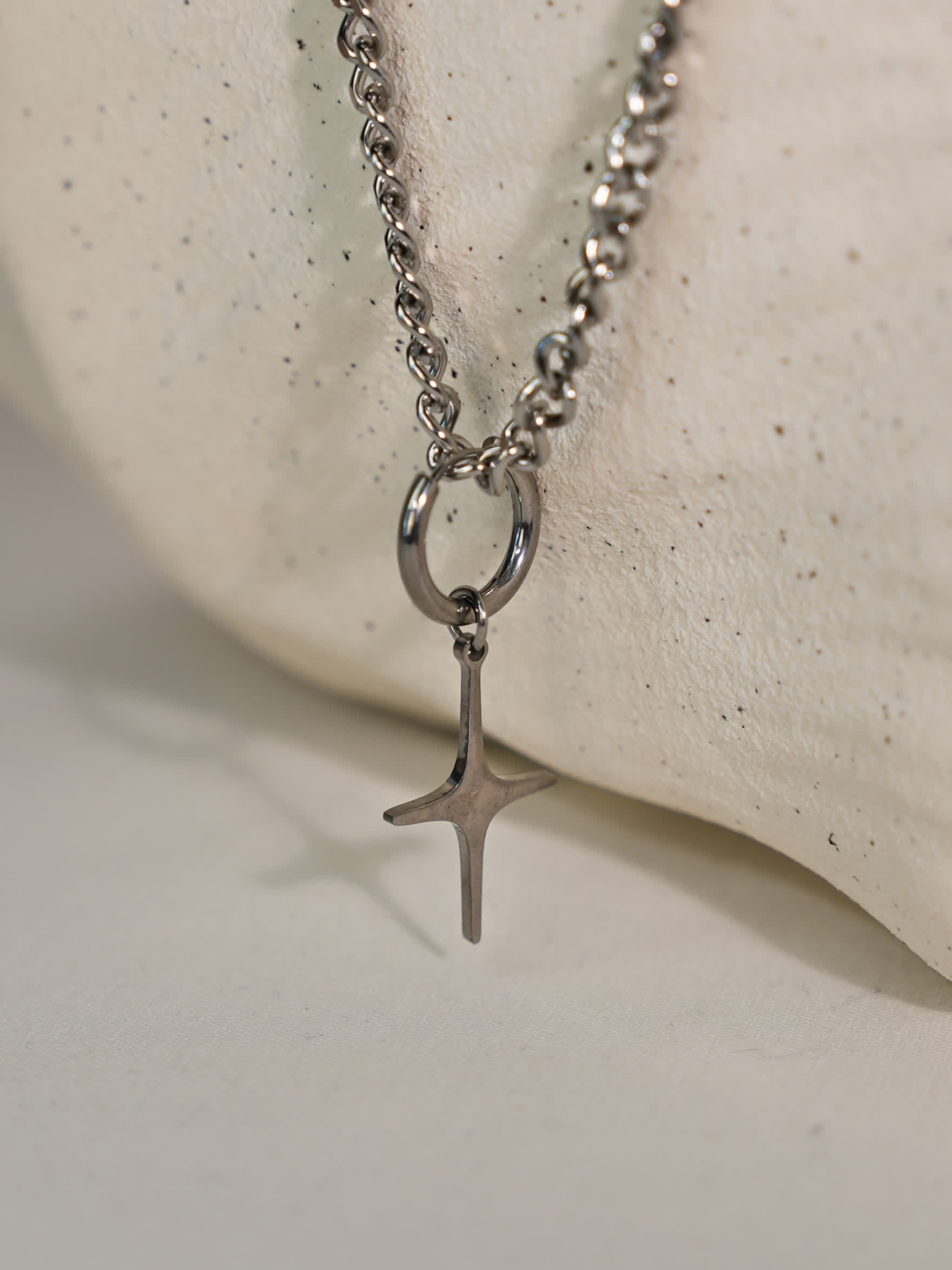 Brushed steel sculptural cross pendant hanging from bold chain, displayed on a curved white stone stand with soft lighting.