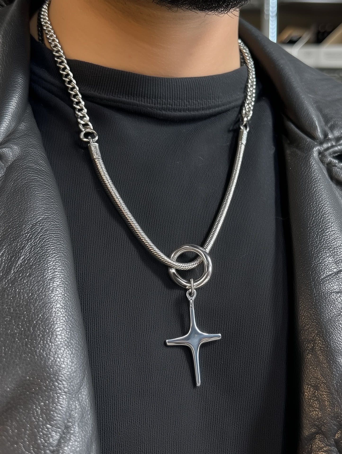 Masculine model wearing brushed stainless steel sculptural cross pendant with bold chain and circular bail, styled with a black shirt and leather jacket.