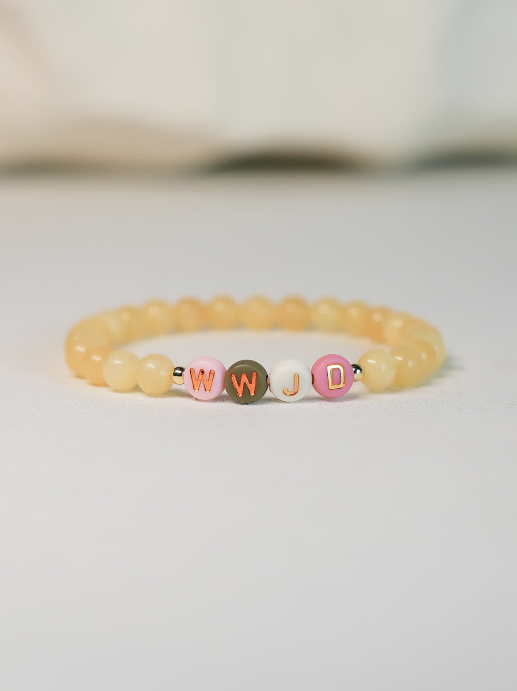 Front-facing image of the WWJD Bracelet on a soft white surface, showing warm yellow gemstone beads and multicolored WWJD letter beads with gold accents.