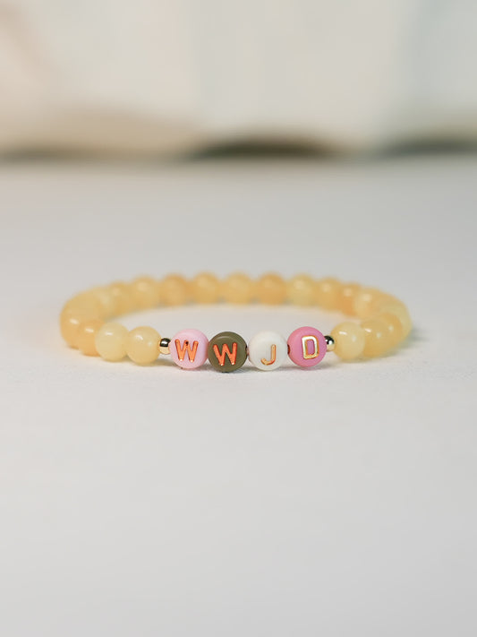 Front-facing image of the WWJD Bracelet on a soft white surface, showing warm yellow gemstone beads and multicolored WWJD letter beads with gold accents.